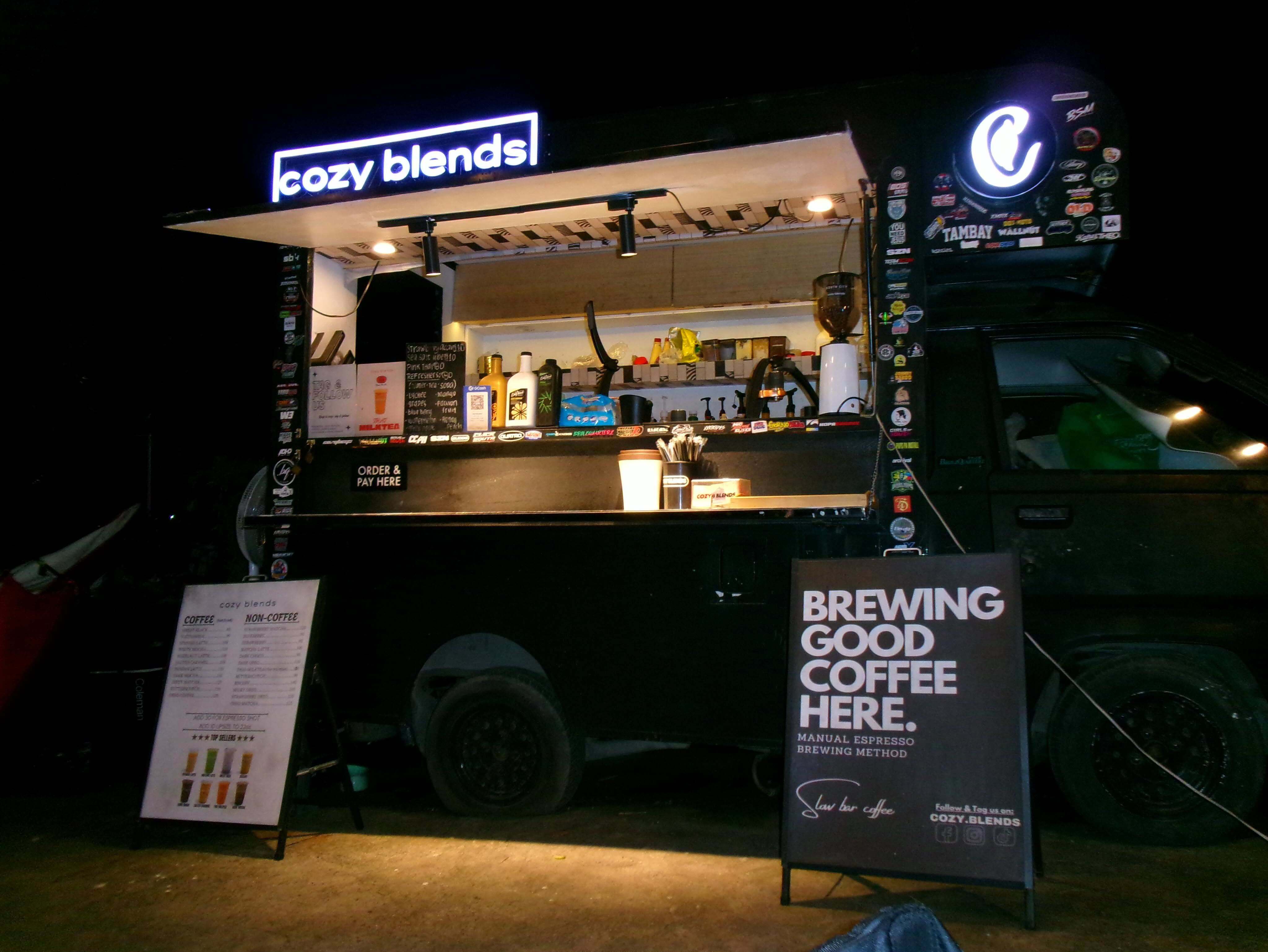 The original cozy blends coffee cart at night, lit by its neon sign, with folding chairs under a tent