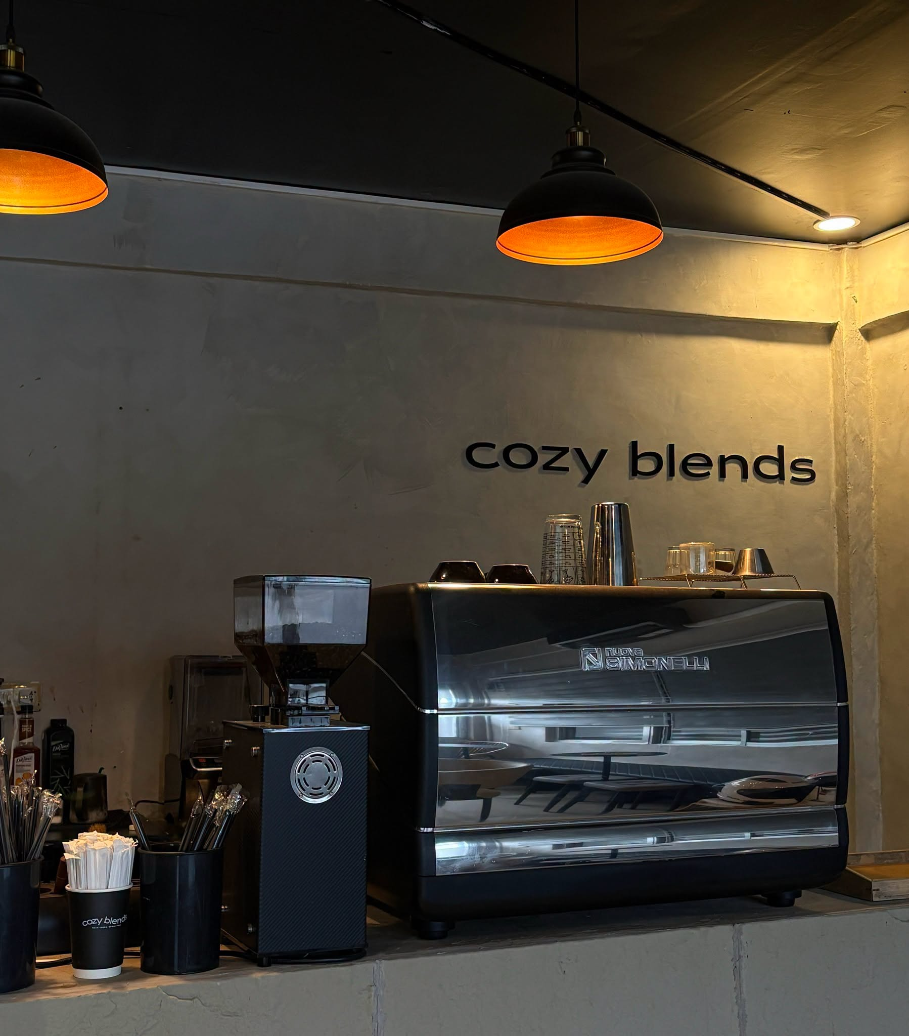Inside the shop: amber pendant lamps over the espresso machine and grinder beneath the cozy blends wall lettering